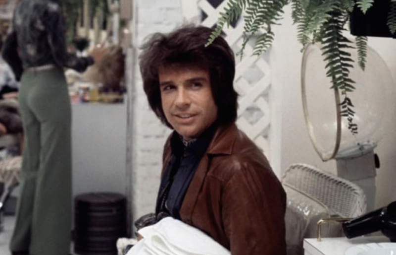 A handsome man in his thirties wearing 1970s-style clothes sits in a hair salon.